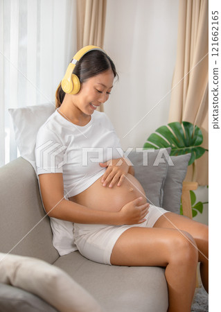 Pregnant woman wearing headphones sitting on couch, smiling and holding her belly, enjoying music in a cozy living room setting. Pregnant woman wearing headphones sitting on couch, smiling and holding her belly, enjoying music in a cozy living room setting. 121662165