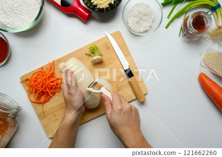 Hands peeling a daikon radish on wooden cutting board surrounded by Kimchi ingredients and spices 121662260