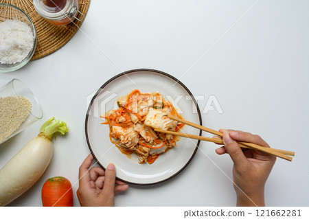 Hand using chopsticks to pick up a piece of kimchi from a plate, surrounded by fresh ingredients Hand using chopsticks to pick up a piece of kimchi from a plate, surrounded by fresh ingredients 121662281