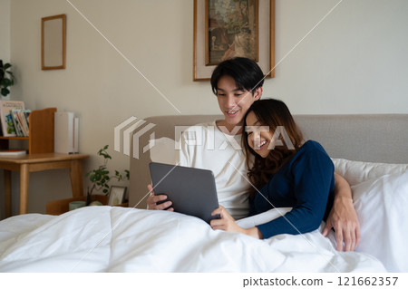 Loving adult couple using digital tablet under warm blanket in a cozy bedroom 121662357