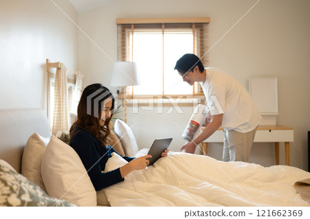 Cheerful woman using digital tablet on bed while her husband vacuuming floor beside Cheerful woman using digital tablet on bed while her husband vacuuming floor beside 121662369