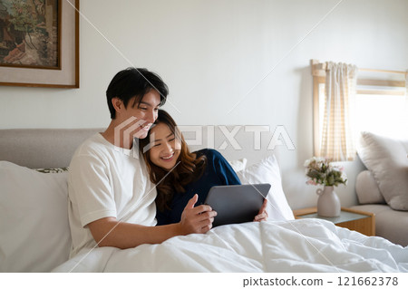 Loving adult couple using digital tablet under warm blanket in a cozy bedroom 121662378