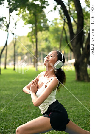 Serene African American woman wearing headphones practicing yoga on mat in a park Serene African American woman wearing headphones practicing yoga on mat in a park 121662422