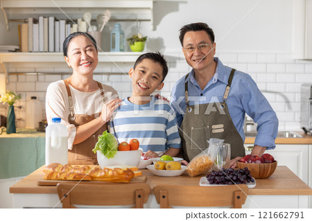 Cheerful Asian family, including grandparents and young grandson stands together in modern kitchen as they prepare meal. Table is filled with fresh ingredients such as vegetables, fruits and bread Cheerful Asian family, including grandparents and young grandson stands together in modern kitchen as they prepare meal. Table is filled with fresh ingredients such as vegetables, fruits and bread 121662791