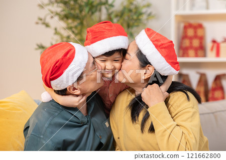 Heartwarming Christmas moment as grandparents, both wearing Santa hats, give their smiling grandchild kiss on cheeks. Child is sandwiched between loving couple, also wearing Santa hat 121662800