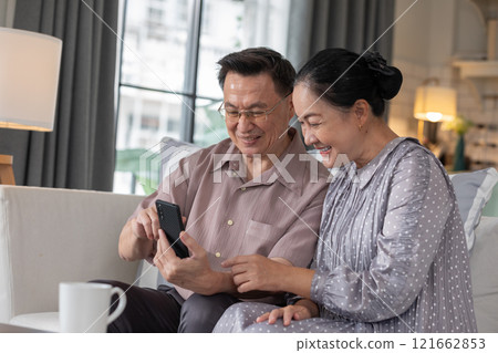 Asian senior couple sitting on couch, happily looking at smartphone screen. They are smiling and sharing moment of connection, showcasing the joy of exploring technology together in cozy home setting Asian senior couple sitting on couch, happily looking at smartphone screen. They are smiling and sharing moment of connection, showcasing the joy of exploring technology together in cozy home setting 121662853
