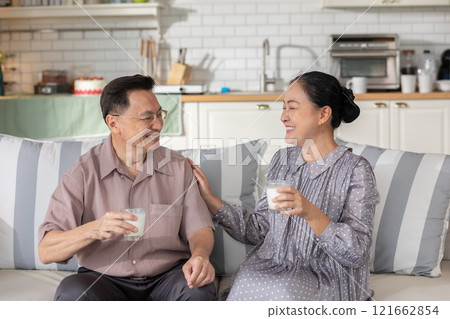 Joyful scene of Asian senior couple sitting on couch at home, smiling and holding glasses of milk. They share warm and affectionate moment, showcasing love and companionship in cozy kitchen setting Joyful scene of Asian senior couple sitting on couch at home, smiling and holding glasses of milk. They share warm and affectionate moment, showcasing love and companionship in cozy kitchen setting 121662854