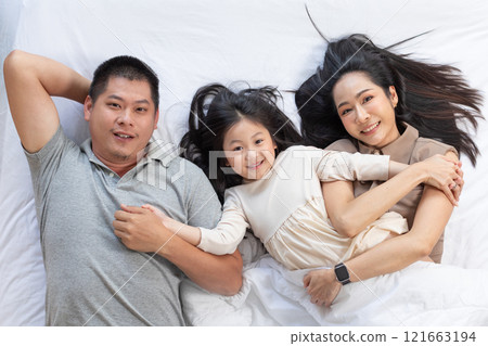 Cheerful Asian family scene featuring father, mother and daughter lying together on bed. They are all smiling and looking at camera, capturing warm and loving family moment 121663194