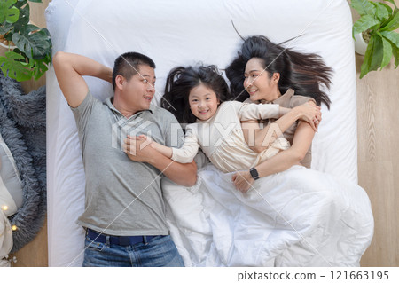 Asian joyful family scene with father, mother, and daughter lying together on bed. They are smiling and looking at each other, sharing moment of closeness and warmth 121663195
