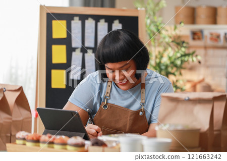 smiling entrepreneur wearing apron writes notes preparing food delivery orders in modern, well-lit workspace. includes tablet, packaging bags indicating small business or home-based food delivery smiling entrepreneur wearing apron writes notes preparing food delivery orders in modern, well-lit workspace. includes tablet, packaging bags indicating small business or home-based food delivery 121663242