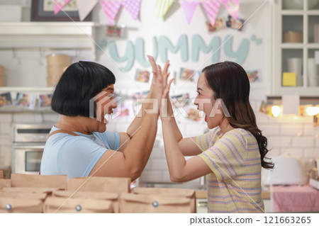 Two women enthusiastically high-fiving in vibrant and creative kitchen, celebrating moment of success or achievement. emphasizing joyful and collaborative atmosphere. teamwork, partnership concept 121663265