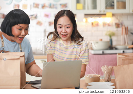 Two cheerful Asian chefs share a joyful moment as they look at a laptop in a cozy kitchen. Surrounded by takeaway bags and delicious food items, they exhibit a sense of accomplishment and happiness 121663266