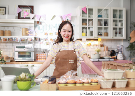 confident woman entrepreneur stands proudly in her kitchen, surrounded by prepared food items neatly packaged for sale. She wearing apron and smiling, exuding sense of accomplishment and professional 121663286
