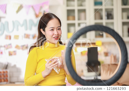 Female vlogger in bright yellow sweater enthusiastically presents food product in cozy kitchen. Captured mid-presentation through ring light, with smartphone positioned in front of her for recording Female vlogger in bright yellow sweater enthusiastically presents food product in cozy kitchen. Captured mid-presentation through ring light, with smartphone positioned in front of her for recording 121663293