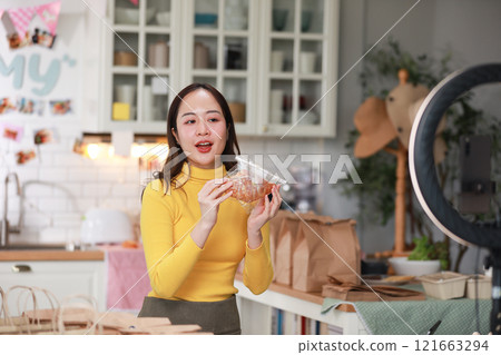 Female vlogger in bright yellow sweater enthusiastically presents food product in cozy kitchen. Captured mid-presentation through ring light, with smartphone positioned in front of her for recording 121663294
