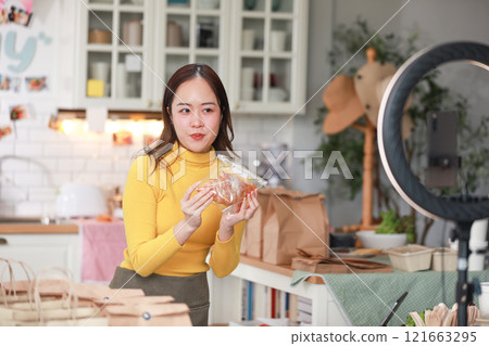 Female vlogger in bright yellow sweater enthusiastically presents food product in cozy kitchen. Captured mid-presentation through ring light, with smartphone positioned in front of her for recording 121663295