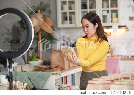 Female vlogger in bright yellow sweater enthusiastically presents food product in cozy kitchen. Captured mid-presentation through ring light, with smartphone positioned in front of her for recording 121663296