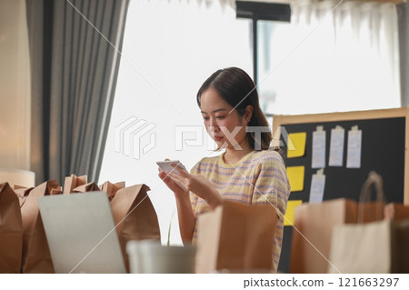 Focused Asian woman carefully managing food delivery orders in well-organized workspace. Surrounded by paper bags and working with notes. business management concept Focused Asian woman carefully managing food delivery orders in well-organized workspace. Surrounded by paper bags and working with notes. business management concept 121663297