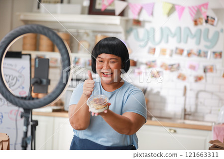 Enthusiastic Asian Content Creator Presenting Food Item in Modern Kitchen Setup, including ring light and smartphone, to film culinary tutorial or review. Small Business Concept Enthusiastic Asian Content Creator Presenting Food Item in Modern Kitchen Setup, including ring light and smartphone, to film culinary tutorial or review. Small Business Concept 121663311