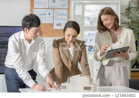 Asian business group of professionals working together on an architectural project in a modern office. The team is engaged in reviewing a physical model, with one person taking notes on a tablet 121663802