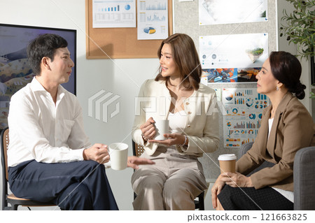 Asian group of business professionals enjoying casual conversation during coffee break in modern office. The trio holding coffee cups engages in relaxed discussion. 121663825