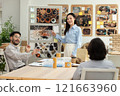 Asian business team engaged in creative discussion during presentation in modern office. Woman points to visual board fill with images and notes as her colleagues actively participate in conversation 121663960
