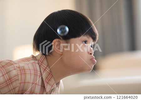 Asian cheerful young boy with Down syndrome enjoys playing with bubbles indoors. His bright smile and playful gestures capture the innocence and joy of childhood 121664020