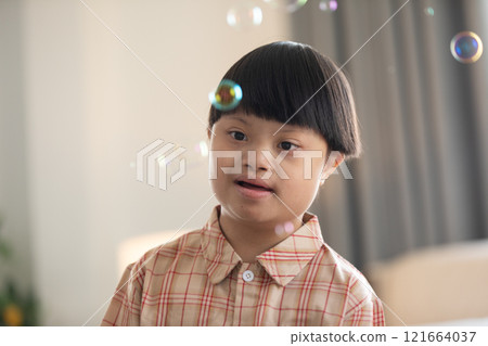 Asian cheerful young boy with Down syndrome enjoys playing with bubbles indoors. His bright smile and playful gestures capture the innocence and joy of childhood Asian cheerful young boy with Down syndrome enjoys playing with bubbles indoors. His bright smile and playful gestures capture the innocence and joy of childhood 121664037