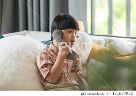 Asian young boy with Down syndrome sits comfortably at home, engaged in a phone conversation. He holds a retro-style telephone, focusing intently on the call Asian young boy with Down syndrome sits comfortably at home, engaged in a phone conversation. He holds a retro-style telephone, focusing intently on the call 121664038