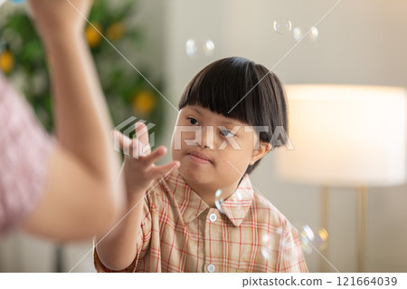 Asian cheerful young boy with Down syndrome enjoys playing with bubbles indoors. His bright smile and playful gestures capture the innocence and joy of childhood 121664039