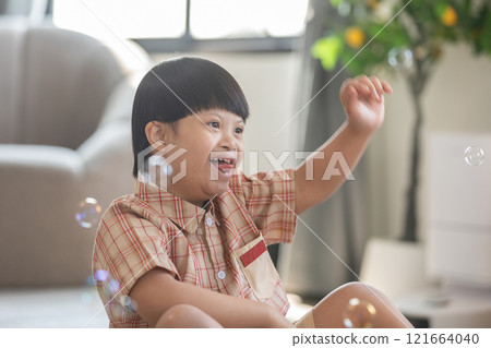 Asian cheerful young boy with Down syndrome enjoys playing with bubbles indoors. His bright smile and playful gestures capture the innocence and joy of childhood Asian cheerful young boy with Down syndrome enjoys playing with bubbles indoors. His bright smile and playful gestures capture the innocence and joy of childhood 121664040