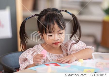 Asian young girl with Down syndrome wearing pigtails, is immersed in arts and crafts project at table. She holds piece of colored paper with focus and concentration, surrounded by crafting materials Asian young girl with Down syndrome wearing pigtails, is immersed in arts and crafts project at table. She holds piece of colored paper with focus and concentration, surrounded by crafting materials 121664054