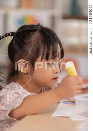 Asian young girl with Down syndrome wearing pigtails, is immersed in arts and crafts project at table. She holds piece of colored paper with focus and concentration, surrounded by crafting materials Asian young girl with Down syndrome wearing pigtails, is immersed in arts and crafts project at table. She holds piece of colored paper with focus and concentration, surrounded by crafting materials 121664055