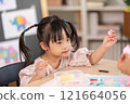 Asian young girl with Down syndrome wearing pigtails, is immersed in arts and crafts project at table. She holds piece of colored paper with focus and concentration, surrounded by crafting materials 121664056