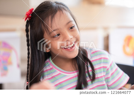 Asian joyful young girl with Down syndrome smiles warmly while engaged in a creative activity. Her cheerful expression highlights the happiness and warmth of the moment 121664077
