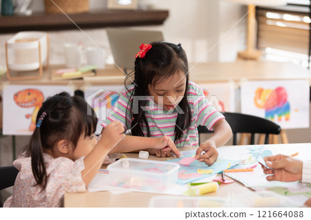 Two young Asian girls with Down syndrome engage in a fun arts and crafts activity, sharing smiles and creativity. They are focused on crafting with colorful materials 121664088