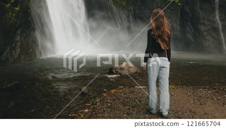 Female hiker is admiring a powerful waterfall cascading down a rocky cliff in a lush tropical rainforest. She is enjoying the peace and serenity of nature during her adventure travel in indonesia 121665704
