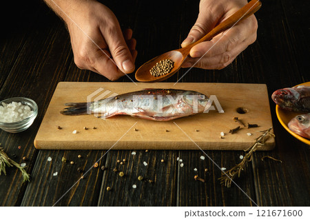 Hands gently sprinkle spices over a freshly caught fish laid out on a wooden cutting board. Surrounding herbs and seasonings create an inviting atmosphere in a rustic kitchen. Hands gently sprinkle spices over a freshly caught fish laid out on a wooden cutting board. Surrounding herbs and seasonings create an inviting atmosphere in a rustic kitchen. 121671600
