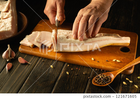 Hands skillfully cut through fresh pork fat on a wooden board, surrounded by garlic cloves and spices. The warm light enhances the rustic feel of this culinary moment, showcasing craftsmanship 121671605