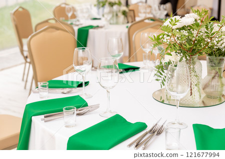 Close up of wine glasses, silver tableware and green napkins on round tables covered with white tablecloth. Close up of wine glasses, silver tableware and green napkins on round tables covered with white tablecloth. 121677994