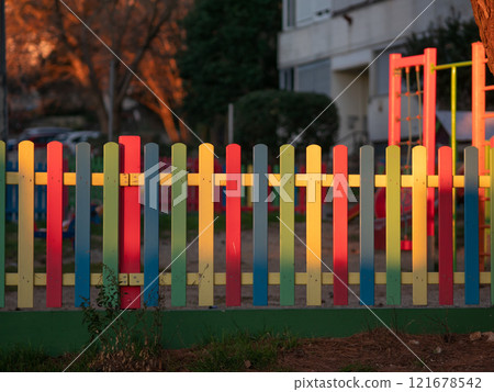 The colorful painted wooden boards on the fence around a playground are illuminated by the rays of the setting sun. Pula, Croatia - January 3, 2025 121678542
