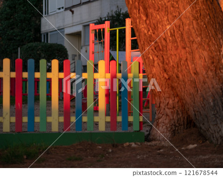 Pine tree illuminated at sunset. Tree trunk. Multicolored painted wooden boards on the fence around the playground are illuminated by the rays of the setting sun. Pula, Croatia - January 3, 2025 Pine tree illuminated at sunset. Tree trunk. Multicolored painted wooden boards on the fence around the playground are illuminated by the rays of the setting sun. Pula, Croatia - January 3, 2025 121678544