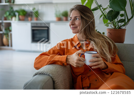 Happy woman smiles with coffee mug in hand on couch, looking around cozy domestic living room alone 121678585