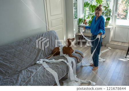 Dog causes humorous chaos, unraveled toilet paper around room, while puzzled female owner reprimands 121678644