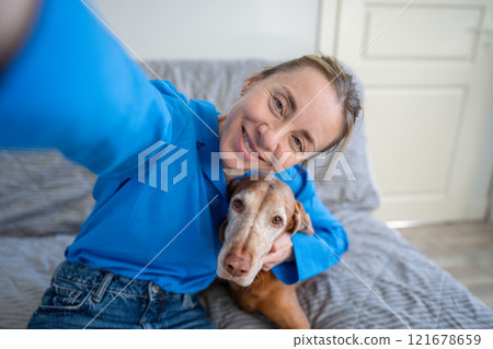 Selfie of smiling woman embracing Vizsla dog on couch at home. Owner affection, pet healthcare, love 121678659