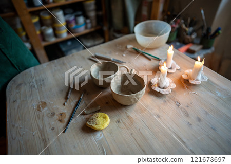Pottery workshop wooden table featuring candles, clay tools, sponge and white clay bowls, cups Pottery workshop wooden table featuring candles, clay tools, sponge and white clay bowls, cups 121678697