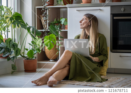 Pensive woman with laptop looking at window, feeling lack of energy sit on floor at home with plants 121678711