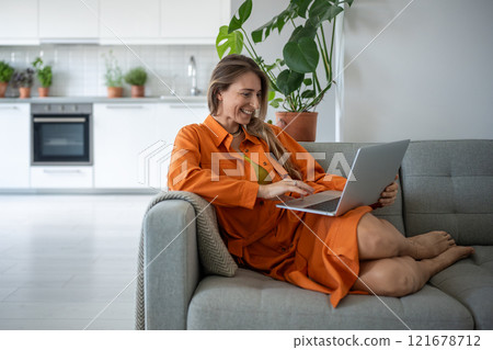 Joyful middle aged woman using laptop smiling relaxing on couch spending leisure time at cozy home. 121678712