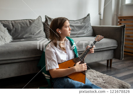 Thoughtful woman in earbuds listens to music while playing balalaika strings on floor in living room 121678826