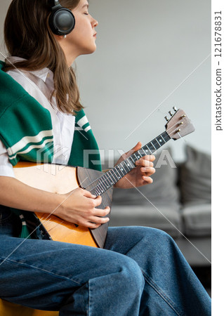 Woman enjoy music, playing balalaika, listening to songs through headphones at home. Ear training 121678831